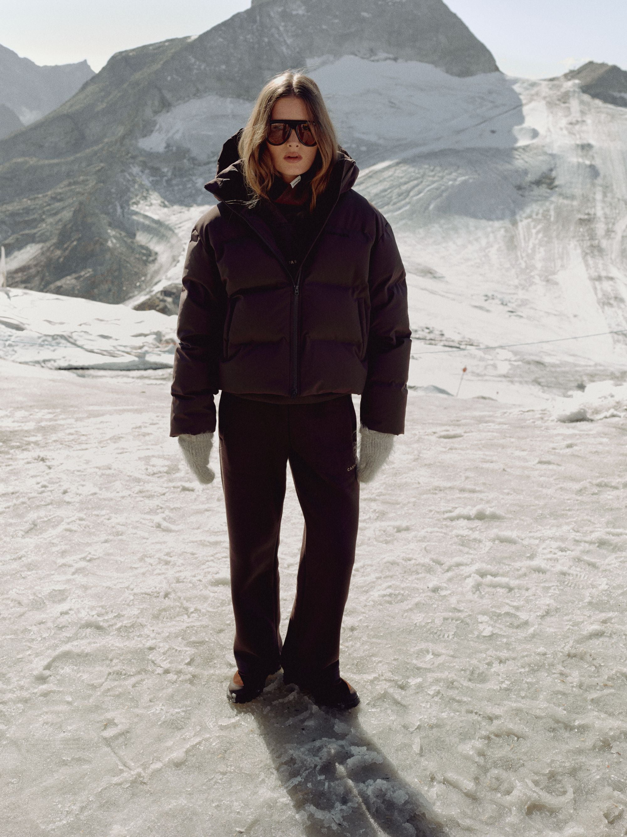 Woman wearing a cocoa bean CASPARA Aurora puffer jacket in a snowy mountain landscape.